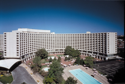 An aerial view of a large modern hotel building with a flat roof. The building is white and has multiple floors and balconies. It appears to be a high-rise apartment complex with multiple levels. In front of the building there is a large swimming pool with lounge chairs and umbrellas scattered around it. The pool is surrounded by trees and greenery and there are several parking lots on the right side of the image. On the left side there are a few other buildings visible in the background. The sky is clear and blue and the overall atmosphere is bright and sunny.