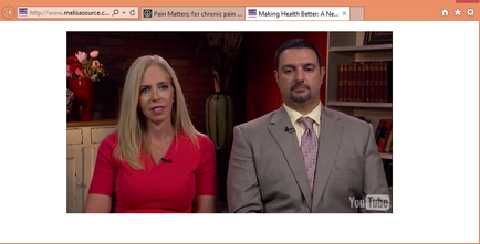 A screenshot of a video interview with a man and a woman. The woman is on the left side of the screen wearing a red dress and has blonde hair. She is looking directly at the camera with a serious expression on her face. The man on the right side is wearing a gray suit and a pink tie. He has a beard and is looking off to the side. In the background there is a bookshelf with books and a vase of flowers. The text on the screen reads "Pain Matters: for chronic pain. Making Health Better. A New."