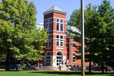 A large brick building with a tall tower on top. The building has multiple windows and arched windows on the front facade. The tower has a pointed roof and is surrounded by trees on both sides. In front of the building there is a grassy area with a few cars parked on the street. There are a few people walking on the sidewalk and a flagpole in the foreground. The sky is blue and the weather appears to be sunny.