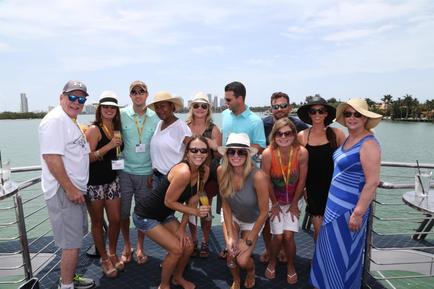 A group of people on a boat posing for a group photo. There are around 20 people in the photo all wearing hats and sunglasses. They are standing on the deck of the boat with the water and buildings visible in the background. The sky is blue and the weather appears to be sunny and warm. Some of the people are smiling and others are looking at the camera. There is a table and chairs on the right side of the image and a railing on the left side.