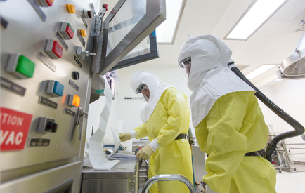 Two people wearing full-body protective suits and helmets in a laboratory setting. They are standing in front of a control panel with various buttons and switches. The person on the left is holding a piece of paper and appears to be working on a machine while the person in the middle is using a vacuum cleaner. The control panel has a red sign that reads "VAC" and there is a large screen above it. The background shows other laboratory equipment and equipment. The overall atmosphere of the image is clean and sterile.