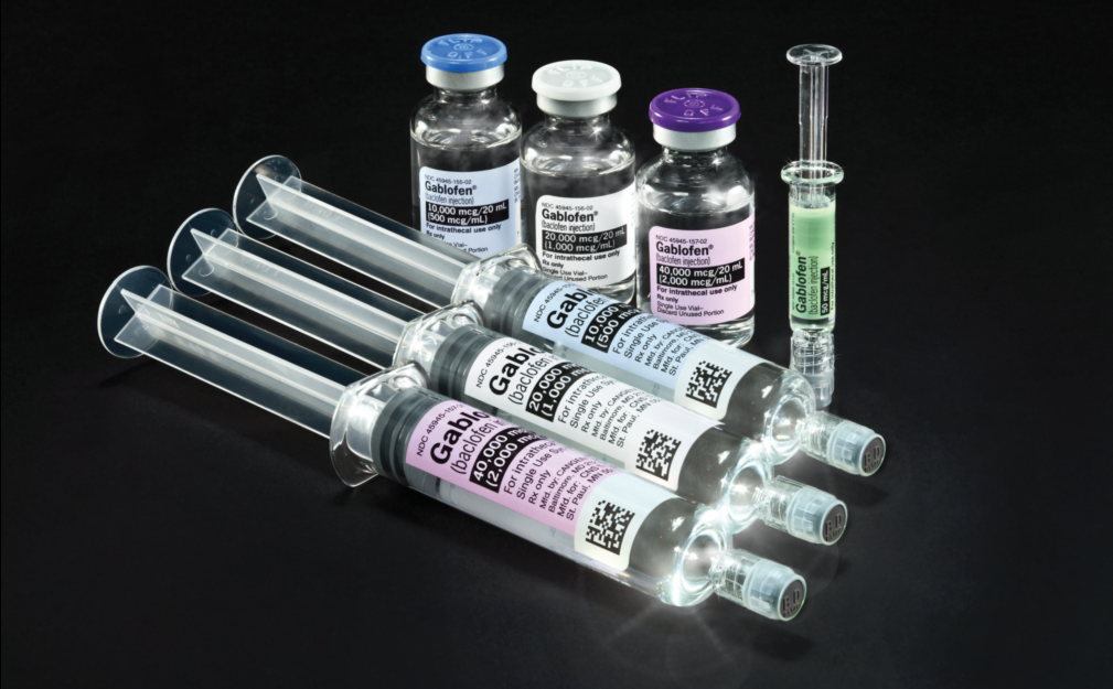 A group of three prefilled syringes of Gablofen (laying flat in the foreground) with thee vials of Gablofen & one syringe of Gablofen (standing vertically in the background) of various dosages (sometimes visible and sometimes partially obscured). Both the syringes and the vials are transparent and all have labels (of various colors) with black text. The lids of the vials color coordinate with their labels (ex: the 40000 mcg/20mL dosage has both a purple label and a purple lid). The background is a black gradient.