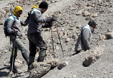 Three men hiking on a rocky terrain. They are all wearing backpacks and carrying trekking poles. The man in the middle is wearing a gray jacket black pants and a yellow beanie while the man on the left is carrying a blue backpack. All three men are wearing sunglasses and appear to be focused on their hike. The terrain is covered in small rocks and boulders and the sky is visible in the background.