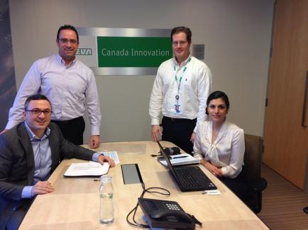 Four people in an office setting. There is a desk in the center of the image with a laptop a phone and a water bottle on it. On the left side of the desk there is a man sitting and smiling at the camera. Next to him there are two men and a woman. The man on the left is wearing a blue shirt and glasses the man in the middle is standing and smiling and the woman on the right is sitting on a chair. Behind them two men are standing and posing for the photo. The background shows a wall with a sign that reads "Canada Innovation".