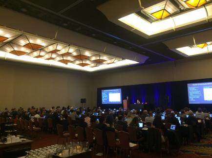 A large conference room with a high ceiling and a chandelier hanging from it. The room is filled with people sitting in rows of chairs facing a stage where a man is giving a presentation. The stage is covered with a blue curtain and there are two large screens on either side of the stage. The people in the room appear to be engaged in the presentation with some sitting at tables and others standing at the front of the room. The tables are covered with white tablecloths and have glasses and plates on them. The floor is carpeted and there is a large window on the left side.