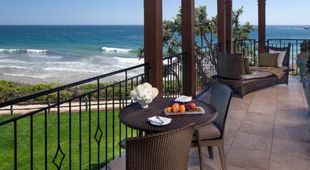 A beautiful balcony overlooking the ocean. The balcony has a tiled floor and a black railing. There is a round table with a vase of white flowers and a plate of fruit on it. There are two wicker chairs and a sofa on the balcony providing a comfortable seating area. The ocean is visible in the background with waves crashing onto the shore. The sky is blue and the weather appears to be sunny and warm.