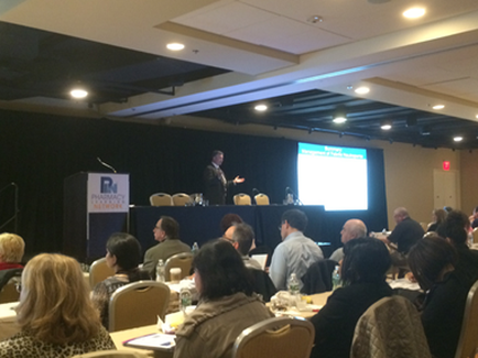 A large conference room with a stage and a projector screen. The room is filled with people sitting at tables facing the stage where a man is giving a presentation. The man is standing at a podium and speaking into a microphone. He is wearing a suit and tie and appears to be giving a speech. The audience is attentively listening to the presentation. There is a banner on the left side of the room with the logo of a company on it. The walls are painted in a dark color and there are rows of chairs in the room.