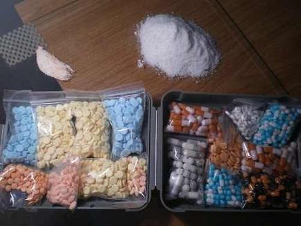 Two plastic containers filled with different types of pills. The containers are placed on a wooden surface. On the left side of the image there is a small pile of white powder which appears to be a type of granular or granular substance. The container on the right side is filled with a variety of pills in different colors and sizes. The pills are arranged in a neat and orderly manner with some of them spilling out of the containers. The colors of the pills range from light blue to dark blue and some have a metallic sheen. There is also a small piece of white foam on the top right corner of the container.