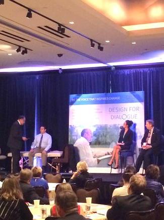 A group of people sitting in a conference room with a large screen in the background. The screen displays an image of a man and a woman dancing on a stage with the text "Design for Dialogue" above them. The room is dimly lit with blue and purple lights and there are rows of chairs on either side of the screen. The people in the room appear to be engaged in a conversation with some sitting at tables and others standing.
