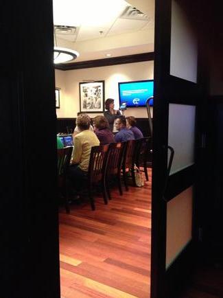 A group of people sitting on wooden chairs in a room with a wooden floor. The room appears to be a conference room or a meeting room as there is a large screen on the wall in the background. The people are gathered around a table and there are several framed pictures hanging on the walls. The image is taken from a high angle looking down on the people and the table. The overall mood of the image is casual and relaxed.