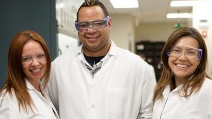 Three people two women and a man standing in a laboratory. They are all wearing white lab coats and safety goggles. The man is in the middle with the two women on either side of him. All three are smiling and appear to be happy. The background is blurred but it appears to be a clean and well-lit room with shelves and cabinets.