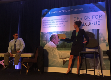 A stage with three people on it. On the left side of the stage there is a man sitting on a chair and a woman standing in front of him. The man is wearing a suit and tie and appears to be giving a presentation. The woman is holding a microphone and is gesturing with her hand as she speaks. Behind them there are two other people sitting on chairs and a large screen with an image of a man and text that reads "Be the voice that inspires change. Design for dialogue." The background is dark and there are blue lights shining on the stage.