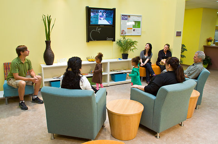 A group of people sitting in a waiting room. There are six people in the room three men and three women all of whom appear to be engaged in a conversation. The room has yellow walls and a large flat-screen TV mounted on the wall. There is a plant in a vase on the left side of the room and a bookshelf on the right side. In the center of the image there is a small table with a child standing in front of it. The people are sitting in blue armchairs and are engaged in conversation.