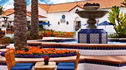A beautiful outdoor courtyard with a fountain in the center. The fountain is made of blue and white tiles and has a birdbath on top. There are two palm trees on either side of the fountain and a small table with a vase of red flowers in front of it. The courtyard is surrounded by colorful flowers and plants and there are several lounge chairs and umbrellas scattered around. In the background there is a white building with a red tiled roof and a sign that reads "Casa del Sol." The sky is blue and the overall atmosphere is peaceful and inviting.