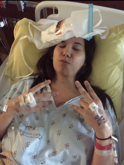 A young woman lying in a hospital bed with her eyes closed. She is wearing a white hospital gown with a floral pattern and has multiple IV tubes attached to her hands. On her head there is a white hat with a blue tube attached to it. The woman appears to be in a state of distress with her head resting on a yellow pillow.