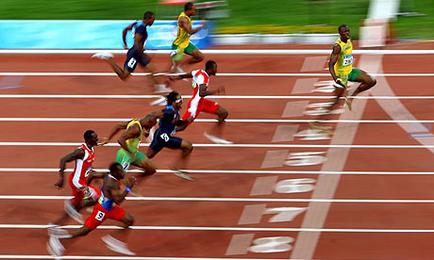 A group of athletes running on a red running track. There are six athletes in the image three of whom are wearing red and white uniforms while the others are wearing yellow and green uniforms. The athletes are in the middle of a race with one athlete in the lead wearing a blue and red uniform and the other two wearing green and yellow uniforms. They are running in a straight line with their arms and legs stretched out in front of them. The background is blurred but it appears to be a stadium with green grass and a blue sky. The image is taken from a high angle looking down on the athletes.