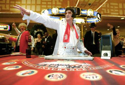 A man in a white suit with a red scarf around his neck and sunglasses. He is standing in front of a red roulette table with the word "Insurance" written on it. The man is pointing upwards with his right hand and has a big smile on his face. Behind him there are several people in a casino some of whom are dancing and cheering. The background is decorated with colorful lights and a large screen. The overall atmosphere appears to be lively and energetic.
