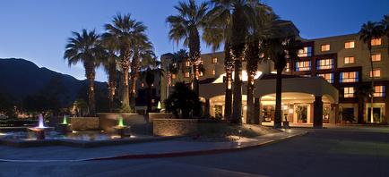 A beautiful hotel at night. The hotel is a two-story building with a beige exterior and large windows. The building is surrounded by palm trees and other tropical plants. In front of the hotel there is a fountain with three water fountains. The sky is dark and the mountains can be seen in the background. The overall atmosphere of the image is peaceful and serene.