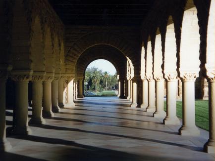 A long arched corridor with multiple columns on either side. The columns are made of stone and have intricate carvings on them. The corridor is made of concrete and has a tiled floor. The ceiling is arched and the columns are supported by columns. In the background there is a view of a park with palm trees and a blue sky. The image is taken from a low angle looking up at the corridor. The overall mood of the image is peaceful and serene.