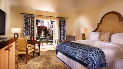 A hotel room with a large king-sized bed in the center. The bed has a wooden headboard and is covered with a blue and white patterned blanket. There are two nightstands on either side of the bed with lamps on them. On the left side there is a desk with a computer monitor and a chair. The room has a large window with blue curtains and a view of a garden outside. The floor is covered in a beige carpet with a floral pattern. The walls are painted in a light yellow color.