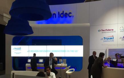 A trade show booth with a blue and white color scheme. The booth has a large screen on the wall with the text "Inidec" and "Tecfidera" written on it. Below the screen there is a banner that reads "2 million infusions administered in the US". There are several people in the booth some standing and some sitting at tables. On the right side of the image there are two large blue pendant lights hanging from the ceiling. The overall atmosphere of the booth is professional and modern.