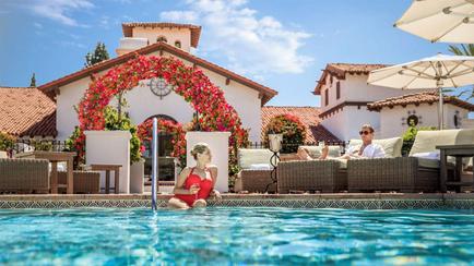 A beautiful outdoor swimming pool with crystal clear blue water. In the center of the image there is a woman in a red swimsuit sitting on the edge of the pool holding a drink in her hand. She appears to be relaxed and enjoying the water. To her right there are two men sitting on lounge chairs one wearing a white shirt and sunglasses and the other wearing a blue shirt. They are both smiling and appear to be enjoying each others company. Behind them is a large white building with a red roof and a clock tower. The building is decorated with pink and red flowers and greenery. There are also several outdoor umbrellas and tables scattered around the pool area. The sky is blue and there are palm trees in the background.