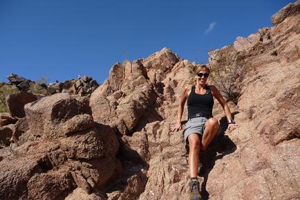 A young woman sitting on a large rock formation. She is wearing a black tank top gray shorts and black hiking boots. She has blonde hair and is wearing sunglasses. The rock formation is made up of large boulders and there are other rocks and boulders scattered around. The sky is blue and the weather appears to be clear and sunny. The woman is smiling and looking off into the distance.
