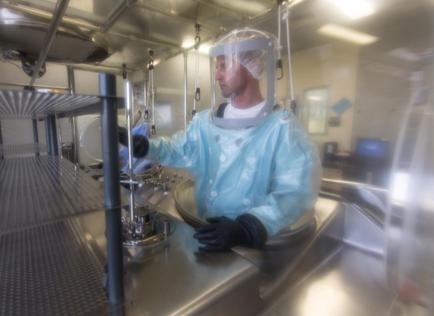 A man wearing a blue protective suit and a white helmet with a visor. He is standing in a large industrial space with stainless steel equipment and racks in the background. The man is wearing black gloves and appears to be working in a laboratory or laboratory setting. He has a serious expression on his face and is looking towards the right side of the image. The image is taken from a low angle looking up at the man and the equipment.