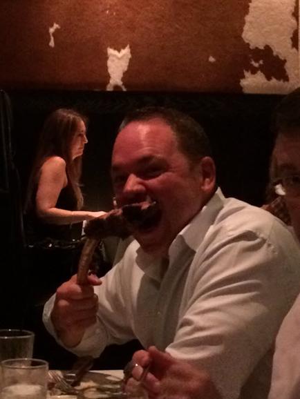 A man sitting at a table in a restaurant. He is wearing a white shirt and is holding a piece of meat in his mouth. He appears to be eating it with a fork and knife. There are other people sitting around the table and a woman is standing in the background. The table is covered with a white tablecloth and there are glasses and plates on it. The walls of the restaurant are decorated with a cowhide pattern.