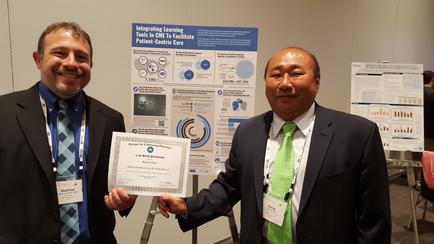 Two men standing next to each other in front of a poster. The man on the left is holding a certificate in his hand and is smiling at the camera. He is wearing a suit and tie and has a name tag around his neck. The poster on the right is a blue and white poster with various graphs and charts on it. The title of the poster is "Integrating Learning Tools to Improve Patient-Centric Care". The poster appears to be a presentation slide with information about the topic of integrating learning tools to improve patient-centric care. The background of the image is a white wall with other posters on display.