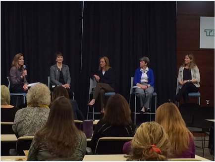 A group of five women sitting on a stage in front of a black curtain. They are all facing the audience and appear to be engaged in a panel discussion. The women are of different ages and ethnicity's and they are all dressed in formal attire. The audience is seated in rows of chairs and there is a projector screen in the background. The image appears to be taken at a conference or presentation.