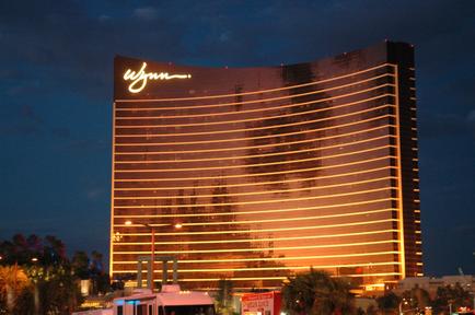 The Wynn Hotel and Casino in Las Vegas Nevada. The hotel is a tall modern building with a curved shape and a large sign that reads "Wynn" in white letters. The building is lit up with orange and yellow lights creating a warm glow. The sky is dark indicating that it is either night or early morning. In front of the hotel there are palm trees and other buildings and a few people can be seen walking around. The overall atmosphere of the image is peaceful and serene.