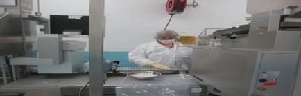 A person wearing a white lab coat a face mask and gloves working in a laboratory. The person is standing in front of a large machine with a red hose attached to it. The machine appears to be a conveyor belt with a bowl of white powder on it. There are other machines and equipment in the background suggesting that the person is working in the laboratory.