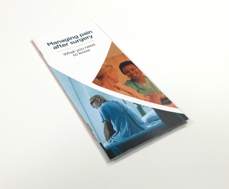 A photograph of a brochure titled "Managing pain after surgery: What you need to know". The cover of the brochure has a white background. Below the title there are two images. <br /><br />The first image on the center right shows two women likely healthcare providers smiling and talking. The photograph has an orange tint. The second image at the bottom left corner shows a man sitting on a hospital bed. He is wearing glasses and a hospital gown and has a serious expression on his face. The second image has a blue tint. <br /><br />The text on the cover is in black and white and is in a modern font. The overall design is simple and professional.