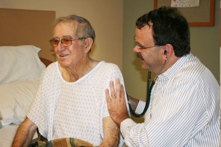 An elderly man sitting in a hospital bed with a nurse standing next to him. The man is wearing a white hospital gown with small white polka dots and glasses. He is smiling and appears to be happy. The nurse is holding a stethoscope around the man's neck and is examining his hand. The background of the image shows a wall with a bulletin board and a window.