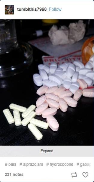 A pile of white and pink pills scattered on a black surface. The pills are of different sizes and shapes with some being round and others being oval-shaped. There is a glass of water in the top left corner of the image and a piece of paper with the word "expand" written on it in the bottom right corner. The background is blurred but it appears to be a table or countertype.