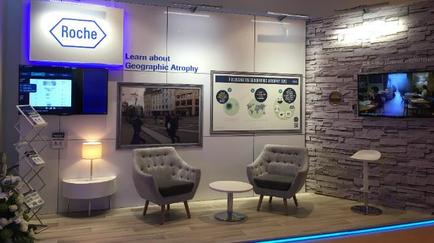 The interior of a booth at a trade show or exhibition. The booth has a white wall with a large screen on the left side displaying the logo of Roche a company that provides information about geographic atrophy. On the right side of the wall there is a large banner with the company's name and logo in blue and white text. In front of the banner there are two gray armchairs and a small white table with a lamp on it. The floor is made of light-colored wood and there are several plants and flowers scattered around the booth. The overall atmosphere of the booth is modern and professional.
