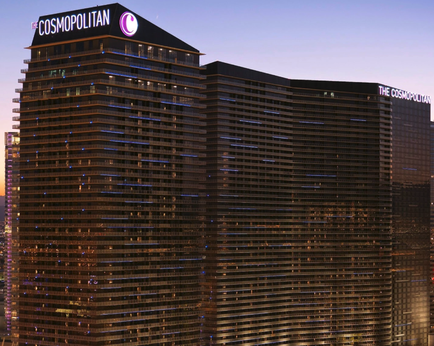 The exterior of the Cosmopolitan Hotel and Casino in Las Vegas Nevada. The hotel is a tall modern building with a black exterior and a pink logo on the top. The building is lit up with blue and purple lights creating a beautiful contrast against the dark sky. In the background there are other buildings and skyscrapers visible. The image appears to be taken at night as the sky is dark and the buildings are lit up.