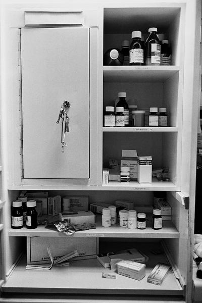 A black and white photograph of an open medicine cabinet. The cabinet is made of metal and has three shelves. On the top shelf there are several bottles of different sizes and colors including black white and red. The bottles are arranged neatly on the shelves and there are also a few small boxes and containers scattered around the bottom shelf. The bottom shelf is empty and the cabinet appears to be in a room with other medical equipment visible in the background.