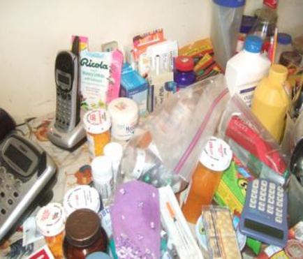 A cluttered desk with various items scattered on it. On the left side of the desk there is a silver telephone and a black cordless phone. Next to the phone there are several bottles of different sizes and colors including blue yellow and pink. In the center of the image there appears to be a plastic bag filled with pills capsules and other medical supplies. The items are spilling out of the bag and onto the desk. The background is a white wall.