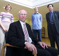 A group of four people standing in a room with a high ceiling and wooden flooring. On the left side of the image there is a woman wearing a pink and white striped shirt and beige pants. She is standing next to a man wearing a black suit and tie. In the center there are two men wearing blue shirts and black suits. The man in the middle is sitting in a wooden chair with his hands resting on his lap. He is wearing glasses and a watch on his left wrist. The other two men are standing behind him also wearing suits and ties. They are all looking at the camera and appear to be posing for the photo.