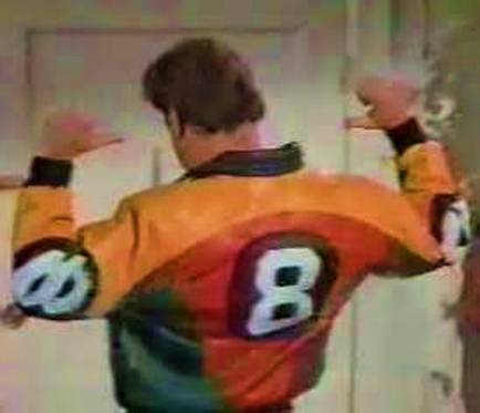 A man standing in a room with his arms stretched out in front of him. He is wearing a colorful jacket with the number 8 on it. The jacket has a unique design with different colors and patterns. The man has short brown hair and appears to be in his late twenties or early thirties. The background is blurred but it seems to be a hallway or a room.
