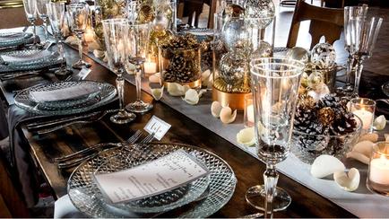 A long table set up for a formal event. The table is covered with a white tablecloth and has a wooden table runner. On the table there are several glassware including wine glasses plates and cutlery. There are also small vases with gold and silver decorations as well as small white flowers and candles. The centerpiece of the table is a large glass vase filled with pine cones and other decorative items. There is also a small white card with a menu on it. The overall color scheme of the setting is silver and gold giving it a luxurious and elegant feel.