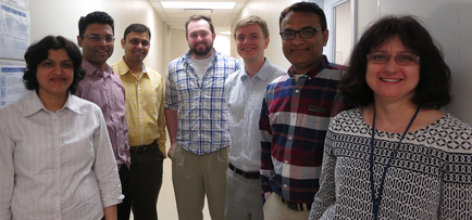 A group of seven people standing in a hallway. They are all smiling and appear to be happy and relaxed. On the left side of the image there is a woman with short dark hair and glasses wearing a white blouse. Next to her there are two men one wearing a blue plaid shirt and the other wearing a yellow shirt. Behind them two men are standing with their arms around each other while the third man is standing with his hands in his pockets. In the background there appears to be a bulletin board with papers pinned to it. The hallway has white walls and a window on the right side.