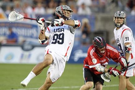 A lacrosse game in progress. The player in the foreground is wearing a white jersey with the number 99 on it and a silver helmet. He is holding a white lacrosse stick and is in the process of making a move towards the goal. Behind him there are two players from the opposing team one wearing a red jersey and the other wearing a gray jersey. The background shows a crowd of spectators in the stands and advertisements on the field. The image appears to have been taken during the day.
