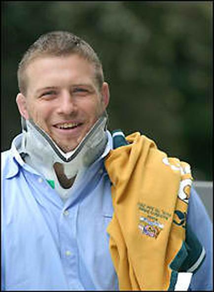 A portrait of a young man with short blonde hair and a big smile on his face. He is wearing a light blue collared shirt and a white neck brace with a green and white logo on it. The man is holding a yellow jacket with a yellow and green design on it which appears to be a scarf or a jacket. The background is blurred but it seems to be an outdoor setting with trees and greenery. The overall mood of the image is happy and relaxed.