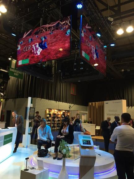A large exhibition hall with a high ceiling and multiple large screens hanging from it. The screens display a video game called "CLR" with a red and blue color scheme. There are several people in the image some standing and some sitting on chairs and a few people walking around the hall. The hall appears to be well-lit with blue and white lights. There is a white counter on the left side of the image with a sign that reads "Medical Information" and a white table with a vase of flowers on it. On the right side there is a podium with a microphone and a display screen. The overall atmosphere of the exhibition hall is busy and bustling.