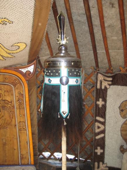 A traditional Mongolian helmet hanging on a wooden pole. The helmet is made of metal and has a silver-colored metal handle with a pointed tip. The body of the helmet is decorated with a blue and white patterned design. The handle is attached to the pole with a long black feather. The background of the image is a tiled wall with a patterned rug hanging on it. On the left side of the wall there is a wooden door with intricate carvings.