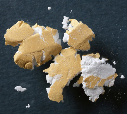 A group of small pieces of yellow and white powder scattered on a black surface. The powder appears to be a type of granular or granular substance with a rough texture and a slightly uneven surface. There are six pieces in total with the largest piece in the center and two smaller pieces on either side. The yellow color of the powder contrasts nicely with the white color creating a striking visual effect. The background is a solid black color making the powder stand out even more.