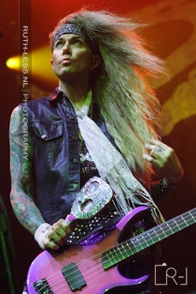 A photograph of a young man playing an electric guitar on stage. He is wearing a black leather vest and has long blonde hair that is blowing in the wind. He has tattoos on his arms and chest and is holding the guitar with both hands. The background is dark and there is a microphone stand visible on the left side of the image. The man appears to be in the middle of a performance as he is looking up at the audience with a focused expression on his face.